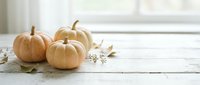 Small white pumpkins and autumn leaves arranged on rustic wooden surface near bright window creating cozy fall seasonal home decor atmosphere.
