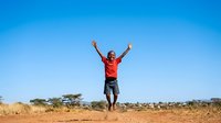 Young Black male child, approximately 8-12 years old, wearing red shirt and dark shorts, jumping with arms raised in celebration on arid sandy terrain under clear blue sky, expressing joy and freedom outdoors.