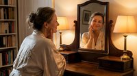 Middle-aged Caucasian woman in white robe sitting at wooden vanity table looking at herself in antique mirror. Warm bedroom interior with bookshelves.