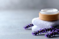 Natural cosmetic cream jar with bamboo lid on white towel, decorated with fresh lavender flowers against light gray background for spa and wellness.