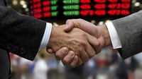 Business handshake between two men in suits against stock market trading board background showing financial data and market trends in red and green numbers.