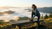 Woman in athletic wear stretching on wooden bench overlooking misty mountain valley at sunrise, outdoor fitness and wellness concept for exercise routines.