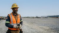 Male construction worker wearing yellow hard hat and orange safety vest standing on industrial construction site with equipment and facilities in background.