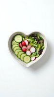 Fresh vegetable salad with cucumber slices, radishes, and mixed greens arranged in a heart-shaped ceramic bowl on white background. Healthy eating concept.