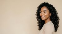 Young Black woman with long curly hair wearing beige ribbed top, smiling at camera against neutral beige background. Portrait for beauty, wellness, and lifestyle content.