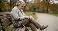 Senior Caucasian woman with gray curly hair sitting on park bench holding walking cane, wearing beige cardigan sweater in autumn outdoor setting.
