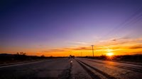 Straight asphalt road stretching toward horizon at golden sunset with power lines, dramatic orange and purple sky, rural landscape at dusk for travel and nature themes.