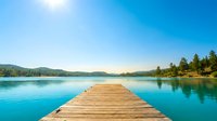 Wooden dock extending into turquoise alpine lake surrounded by evergreen forest and mountains under clear blue sky, suitable for travel and nature backgrounds.