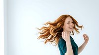 Woman with long wavy auburn hair wearing teal blazer, smiling while looking away against white background. Suitable for business, lifestyle, and professional portraits.