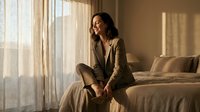 Young professional woman in beige suit sitting on bed by window with natural sunlight streaming through curtains in modern bedroom interior for business lifestyle.
