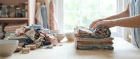 Woman organizing and folding stacked fabric textiles and cloth materials on white table near window for home organization and decluttering project.