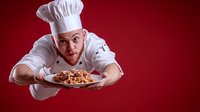 Young male chef in white uniform and hat flying through red background while holding plate of pasta, creative food photography for restaurant advertising.