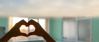 Hands forming heart shape in modern hospital room with soft lighting and medical equipment, symbolizing healthcare compassion and patient care.