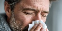 Middle-aged Caucasian man with beard blowing nose into tissue, showing symptoms of cold or flu illness in close-up portrait.
