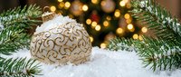 Gold ornament with swirl pattern nestled in snow among evergreen branches and warm bokeh lights, festive holiday decoration for seasonal marketing and winter celebrations.