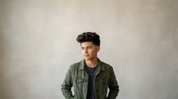 Young man with dark hair and olive skin tone wearing a sage green utility jacket over a black shirt, standing against a neutral gray background. Suitable for lifestyle, fashion, and portrait projects.