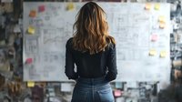 Young woman in black sweater and jeans standing in front of whiteboard with sticky notes and architectural drawings during creative planning session.