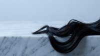 Dark flowing hair strand resting on white marble surface, elegant wave shape against soft gray background, beauty and hair care concept.