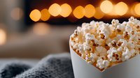 Fresh popcorn overflowing from white bowl against blurred warm bokeh lights background, creating cozy movie night atmosphere. Perfect for entertainment concepts.