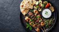 Middle Eastern grilled meat skewers with fresh vegetables, herbs, yogurt sauce and flatbread on dark slate background for Mediterranean cuisine and healthy dining.