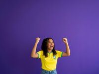 Young woman with long dark hair and medium skin tone wearing bright yellow t-shirt and denim shorts, flexing arms in celebratory pose against vibrant purple background. Suitable for success, achievement, motivation, and empowerment concepts.