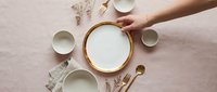 Overhead view of hands arranging empty ceramic bowls and gold utensils on pink linen fabric with dried flowers for table setting or food styling projects.
