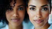 Close up portrait of two young African American women with natural makeup and glowing skin smiling at camera against blurred blue background.