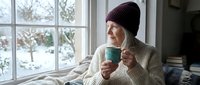 Young woman in cozy knit sweater and beanie enjoying hot drink by window on snowy winter day, perfect for seasonal lifestyle and hygge concepts.