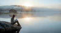 Young man sitting on rocky shore reading book by misty lake at sunrise with wooden cabin in background creating peaceful morning atmosphere.