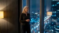 Professional blonde businesswoman in black suit standing by office window overlooking illuminated city skyline at night, corporate executive lifestyle.