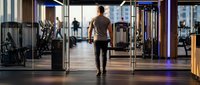 Athletic woman walking through modern fitness center with exercise equipment and large windows creating bright workout environment.