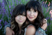 Two young Caucasian sisters with dark wavy hair embracing and smiling in purple lavender field during golden hour summer day for family portrait photography.