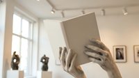 Young woman with medium skin tone holding open book in bright gallery space with natural light from windows, suitable for education, reading, and creative lifestyle content.