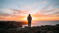 Young adult male in brown jacket standing on rocky coastal cliff overlooking ocean at golden hour sunset, contemplating horizon with dramatic sky and crashing waves below.