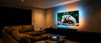 Cozy home theater room with large TV displaying a badger, ambient LED backlight, sectional sofa, and laptop on coffee table in dim lighting.