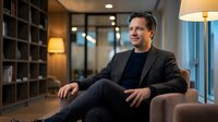 Confident middle-aged Caucasian businessman in dark suit sitting relaxed in modern office lounge, smiling warmly with professional demeanor and positive attitude.