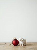Red metallic Christmas ornament ball and white ceramic sheep figurine arranged on wooden surface against neutral background, suitable for holiday greeting cards and seasonal marketing materials.