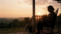 Woman sitting on wooden chair on rural porch at sunset, silhouetted against golden sky and distant mountains, peaceful countryside retreat for relaxation and solitude themes.
