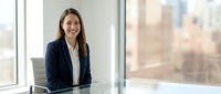 Professional businesswoman smiling at modern office desk near window with city view background for corporate presentations and workplace content.