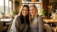 Two happy young women friends embracing and smiling in cozy cafe with warm lighting and plants. Friendship bonding moment for lifestyle content.