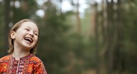 Young girl with light skin tone and blonde hair wearing an orange patterned shirt, laughing joyfully outdoors in a forest setting with tall trees in soft focus background.