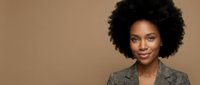 Professional African American businesswoman with natural afro hairstyle wearing gray blazer smiling confidently against neutral beige background.