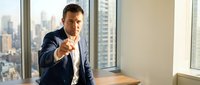 Confident businessman in suit pointing finger at camera in modern office with city skyline view through windows for corporate leadership concepts.