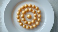 Golden spherical candies arranged in spiral pattern on white ceramic plate, creating minimalist geometric composition with soft natural lighting.