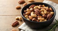 Mixed nuts assortment in black bowl on wooden table with rosemary sprig for healthy snacking and cooking ingredients.