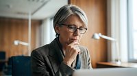 Mature woman with gray hair and glasses wearing olive blazer, hand to chin in thoughtful pose at desk, suitable for business, professional development, leadership, and corporate training materials.