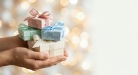Hands holding stack of colorful wrapped gift boxes with ribbons against warm bokeh lights background for holidays and celebrations.