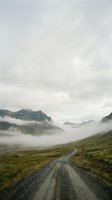 Winding gravel road through misty mountain valley with dramatic peaks shrouded in fog and lush green terrain, suitable for travel and nature backgrounds.