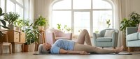 Young woman practicing yoga stretches on floor in bright living room with plants and natural light streaming through large windows.