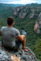 Young athletic man in casual wear sitting on cliff edge overlooking dramatic canyon valley with river below, enjoying peaceful solitary moment in nature.